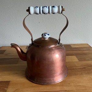 Puede incluir: Una tetera de cobre con un asa y una tapa de porcelana blanca. El asa tiene un diseño floral azul. La tetera está sobre una superficie de madera.