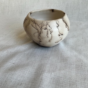 May include: A white ceramic bowl with a unique, hand-painted design of black lines and swirls. The bowl has a rounded shape and a smooth, matte finish.