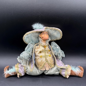 Könnte beinhalten: Eine skurrile Affenpuppe in sitzender Position, gekleidet in aufwendiger Kleidung. Die Puppe trägt einen großen, hellblauen Hut mit goldenen Verzierungen und einer Feder. Das Outfit umfasst eine cremefarbene Weste mit goldenen Akzenten, hellblaue Ärmel und lavendelfarbene Hosen. Die Puppe hat braune Schuhe.