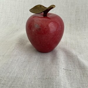 May include: A red marble apple with a gold leaf and stem. The apple is sitting on a white linen surface.