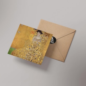 May include: A square greeting card featuring a detail from Gustav Klimt's "Portrait of Adele Bloch-Bauer I" with a gold and yellow color scheme. The card is next to a brown envelope with a black sticker that reads "IN THE WALL".