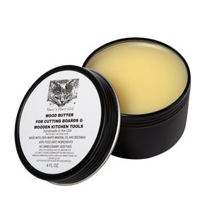 May include: A black metal tin of wood butter for cutting boards and wooden kitchen tools. The tin is open, revealing a yellow, solid balm. The label features a cat illustration and reads "Wood Butter" and "4 FL OZ."