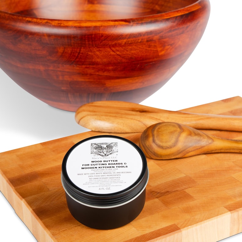 May include: A wooden bowl, cutting board, and spoons are displayed. A black tin of wood butter for cutting boards and wooden kitchen tools is in the foreground. The label reads "WOOD BUTTER FOR CUTTING BOARDS & WOODEN KITCHEN TOOLS".