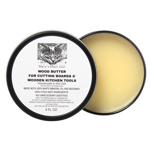 May include: A round tin of wood butter for cutting boards and wooden kitchen tools. The label features a cat illustration and text that reads "Macy's Place LLC" and "4 FL OZ.". The butter has a light yellow color.