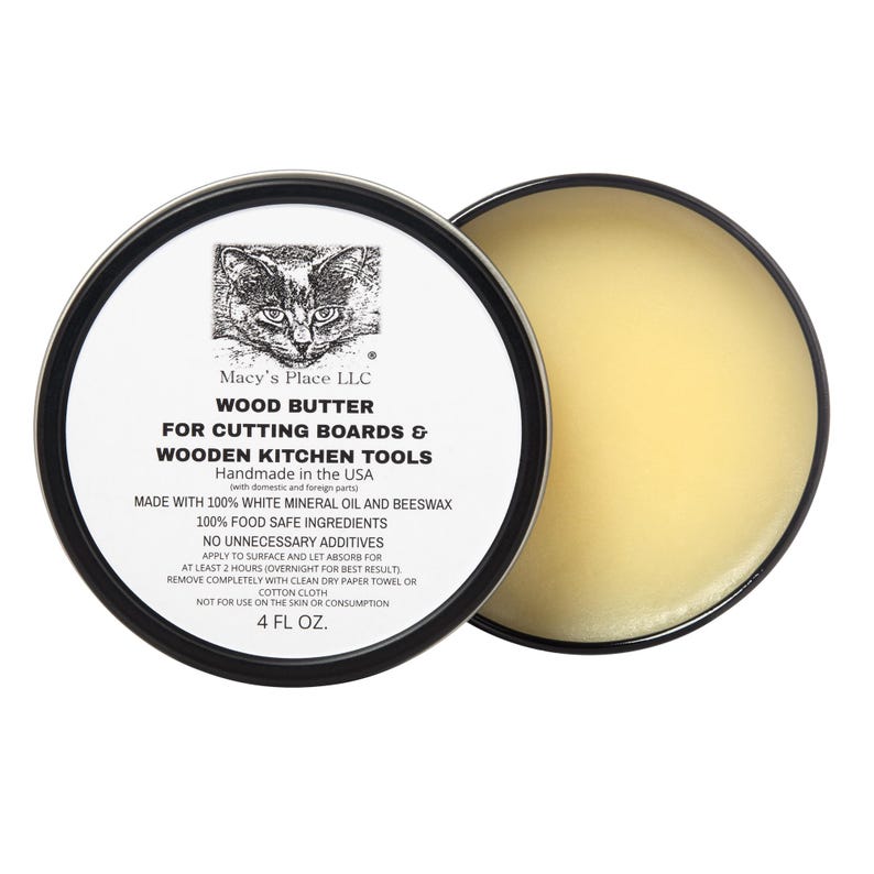 May include: A round tin of wood butter for cutting boards and wooden kitchen tools. The label features a cat illustration and text that reads "Macy's Place LLC" and "4 FL OZ.". The butter has a light yellow color.