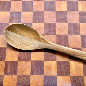 May include: A wooden spoon with a rounded bowl and long handle rests on a checkered cutting board. The spoon is made of light-colored wood, and the cutting board has a pattern of alternating light and dark brown squares.