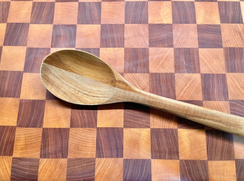 May include: A wooden spoon with a rounded bowl and long handle rests on a checkered cutting board. The spoon is made of light-colored wood, and the cutting board has a pattern of alternating light and dark brown squares.