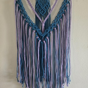 May include: A macrame wall hanging in shades of teal and lavender. The design features a diamond-shaped central element and cascading fringe. The piece is suspended from a natural wooden dowel, adding a touch of rustic charm to the handcrafted textile art.