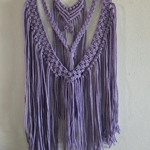 May include: A lavender macrame wall hanging. The handmade piece features intricate knotting patterns, including a central chevron design and cascading fringe. It is suspended from a natural wooden dowel, adding a touch of rustic charm to the decor.