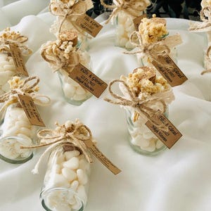 Peut inclure: Petits bocaux en verre remplis de bonbons blancs, d&eacute;cor&eacute;s de fleurs s&eacute;ch&eacute;es, de dentelle et de ficelle. Chaque bocal a un bouchon en li&egrave;ge et une petite &eacute;tiquette avec du texte. Les bocaux sont dispos&eacute;s sur une surface blanche.