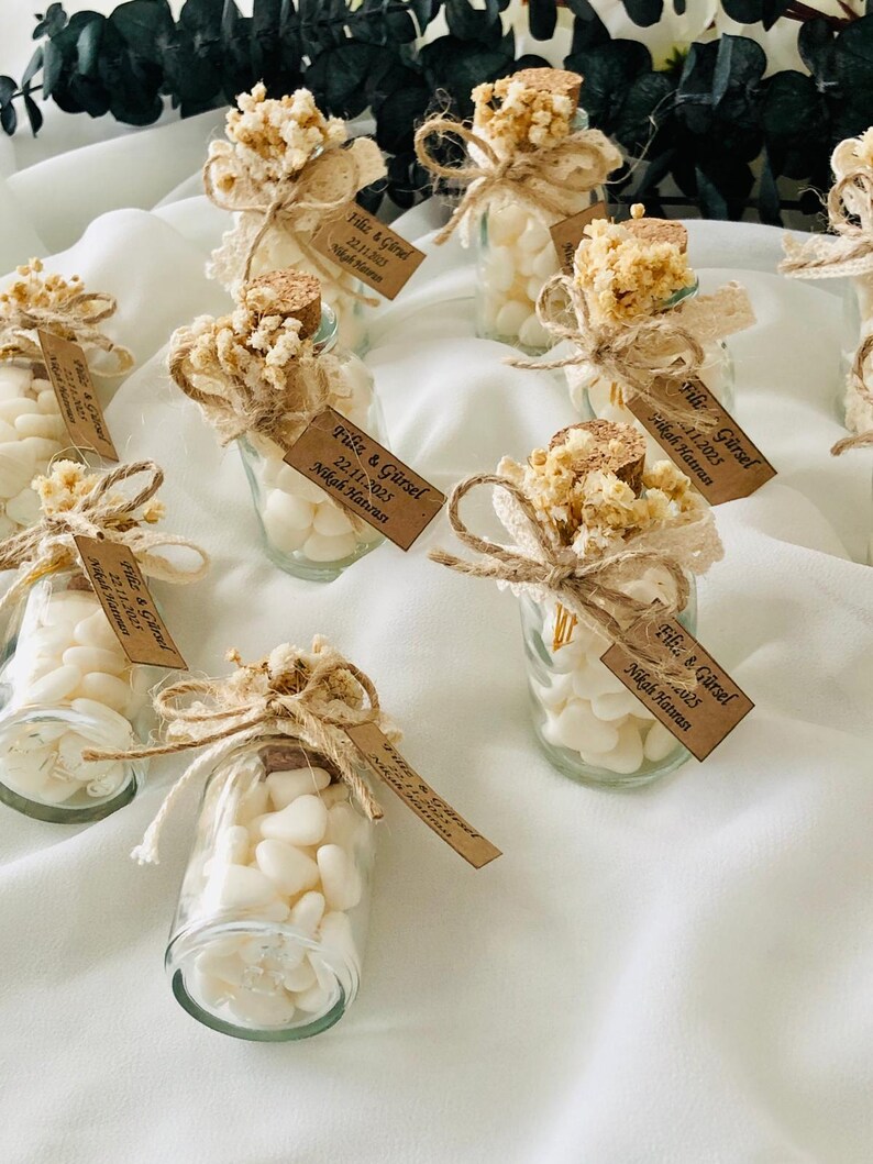 Peut inclure: Petits bocaux en verre remplis de bonbons blancs, d&eacute;cor&eacute;s de fleurs s&eacute;ch&eacute;es, de dentelle et de ficelle. Chaque bocal a un bouchon en li&egrave;ge et une petite &eacute;tiquette avec du texte. Les bocaux sont dispos&eacute;s sur une surface blanche.