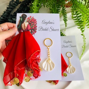 Puede incluir: Recuerdos de despedida de soltera con una bufanda roja con monedas de oro y un llavero de concha blanca. La tarjeta dice "Angelina's Bridal Shower" con una ilustración de una mujer sosteniendo flores. Los recuerdos se exhiben con vegetación en el fondo.