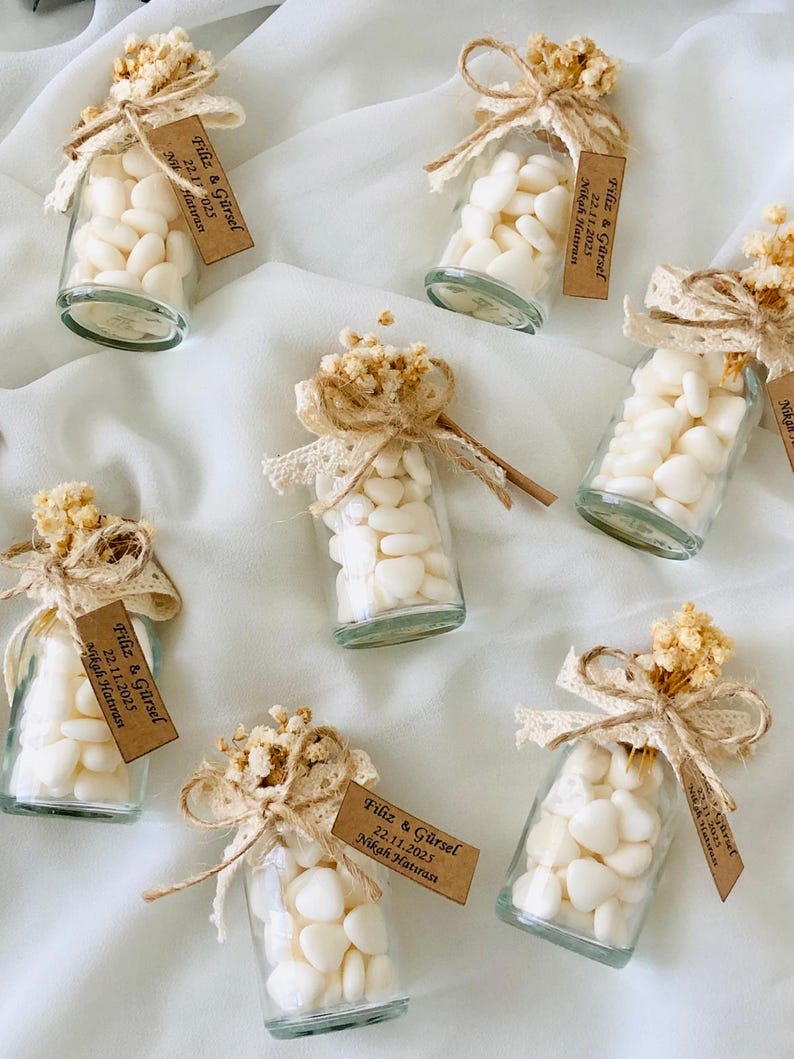 Peut inclure: Petites bouteilles en verre remplies de bonbons blancs en forme de c&oelig;ur, orn&eacute;es de fleurs s&eacute;ch&eacute;es, de dentelle et de ficelle. Chaque bouteille a une petite &eacute;tiquette avec du texte. Une charmante faveur de mariage.