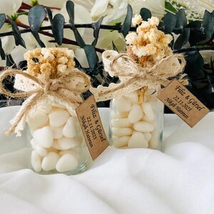 Peut inclure: Deux petits bocaux en verre remplis de bonbons blancs en forme de c&oelig;ur. Chaque bocal est orn&eacute; d'un n&oelig;ud en dentelle et ficelle beige, surmont&eacute; de fleurs s&eacute;ch&eacute;es. De petites &eacute;tiquettes avec du texte sont suspendues aux bocaux.