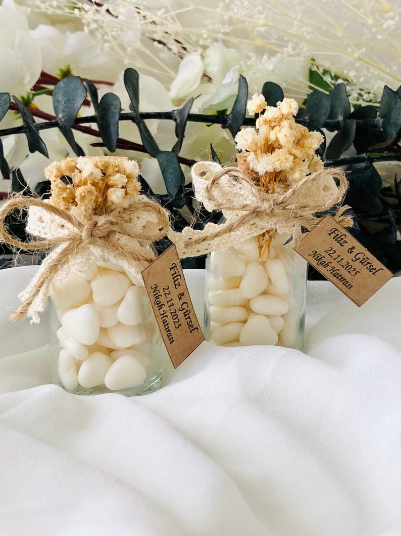 Peut inclure: Deux petits bocaux en verre remplis de bonbons blancs en forme de c&oelig;ur. Chaque bocal est orn&eacute; d'un n&oelig;ud en dentelle et ficelle beige, surmont&eacute; de fleurs s&eacute;ch&eacute;es. De petites &eacute;tiquettes avec du texte sont suspendues aux bocaux.