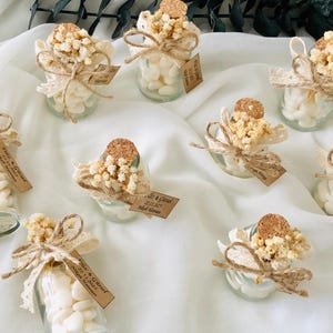 Peut inclure: Petits bocaux en verre remplis de bonbons blancs, d&eacute;cor&eacute;s de dentelle, de ficelle, de fleurs s&eacute;ch&eacute;es et de bouchons en li&egrave;ge. Chaque bocal porte une petite &eacute;tiquette avec du texte. Les bocaux sont dispos&eacute;s sur un tissu blanc.