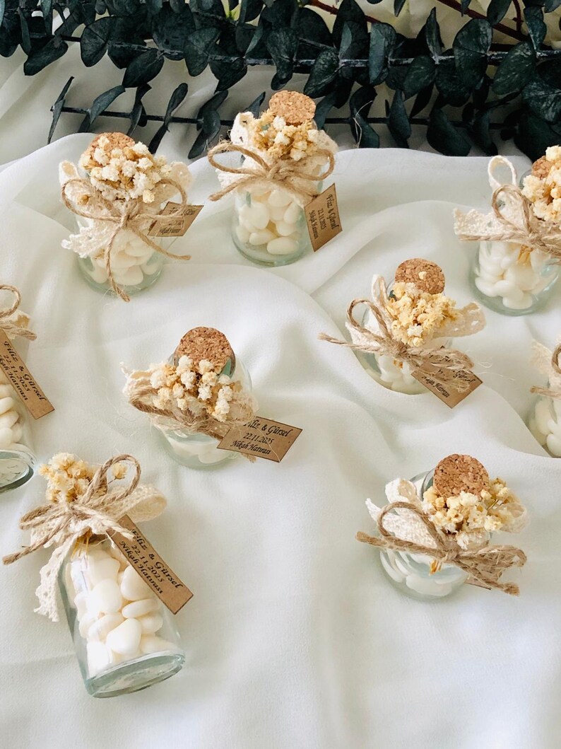 Peut inclure: Petits bocaux en verre remplis de bonbons blancs, d&eacute;cor&eacute;s de dentelle, de ficelle, de fleurs s&eacute;ch&eacute;es et de bouchons en li&egrave;ge. Chaque bocal porte une petite &eacute;tiquette avec du texte. Les bocaux sont dispos&eacute;s sur un tissu blanc.