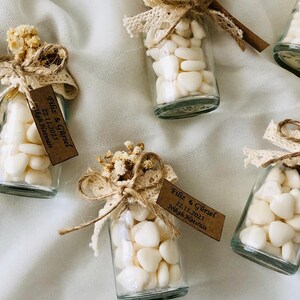 Peut inclure: Petites bouteilles en verre remplies de galets blancs, d&eacute;cor&eacute;es de fleurs s&eacute;ch&eacute;es, de dentelle et de ficelle. Chaque bouteille a une petite &eacute;tiquette marron avec du texte. Les bouteilles sont dispos&eacute;es sur une surface blanche.