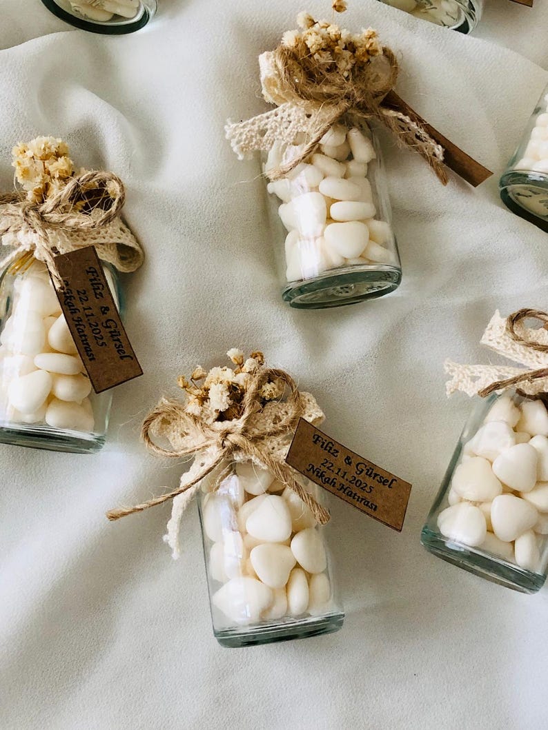 Peut inclure: Petites bouteilles en verre remplies de galets blancs, d&eacute;cor&eacute;es de fleurs s&eacute;ch&eacute;es, de dentelle et de ficelle. Chaque bouteille a une petite &eacute;tiquette marron avec du texte. Les bouteilles sont dispos&eacute;es sur une surface blanche.