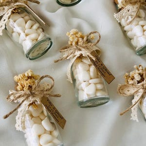 Peut inclure: Petites bouteilles en verre remplies de bonbons blancs, d&eacute;cor&eacute;es de fleurs s&eacute;ch&eacute;es, de dentelle et de ficelle. Chaque bouteille a une petite &eacute;tiquette avec du texte. Les bouteilles sont dispos&eacute;es sur une surface blanche textur&eacute;e.