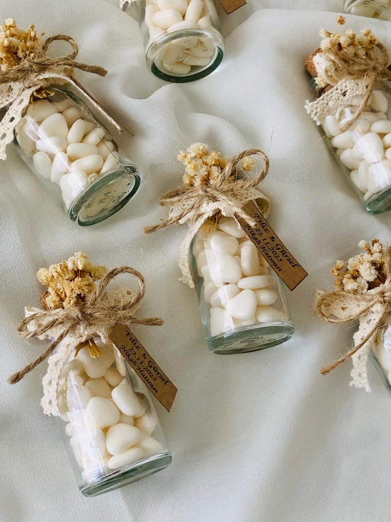 Peut inclure: Petites bouteilles en verre remplies de bonbons blancs, d&eacute;cor&eacute;es de fleurs s&eacute;ch&eacute;es, de dentelle et de ficelle. Chaque bouteille a une petite &eacute;tiquette avec du texte. Les bouteilles sont dispos&eacute;es sur une surface blanche textur&eacute;e.