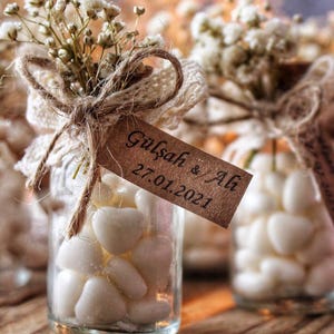 Peut inclure: Petits bocaux en verre remplis de bonbons blancs en forme de c&oelig;ur, attach&eacute;s avec de la ficelle, de la dentelle et des brins de fleurs blanches. Chaque bocal porte une &eacute;tiquette marron avec les noms "G&uuml;lşah & Ali" et la date "27.01.2021".