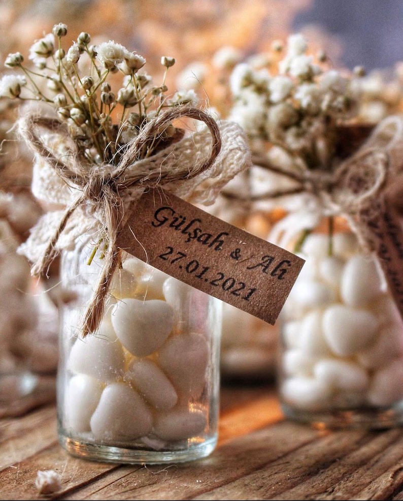 Peut inclure: Petits bocaux en verre remplis de bonbons blancs en forme de c&oelig;ur, attach&eacute;s avec de la ficelle, de la dentelle et des brins de fleurs blanches. Chaque bocal porte une &eacute;tiquette marron avec les noms "G&uuml;lşah & Ali" et la date "27.01.2021".