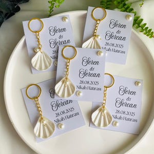May include: A collection of wedding favors featuring white seashell keychains with gold-colored accents and a pearl. Each keychain is attached to a card with the names "Seren & Sercan" and the date "28.08.2025" printed on it.