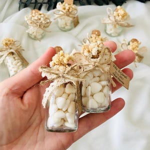 Peut inclure: Petits bocaux en verre remplis de bonbons blancs en forme de c&oelig;ur, orn&eacute;s de fleurs s&eacute;ch&eacute;es, de dentelle et de ficelle. Chaque bocal a un bouchon en li&egrave;ge et une petite &eacute;tiquette. Les bocaux sont dispos&eacute;s sur une surface blanche.