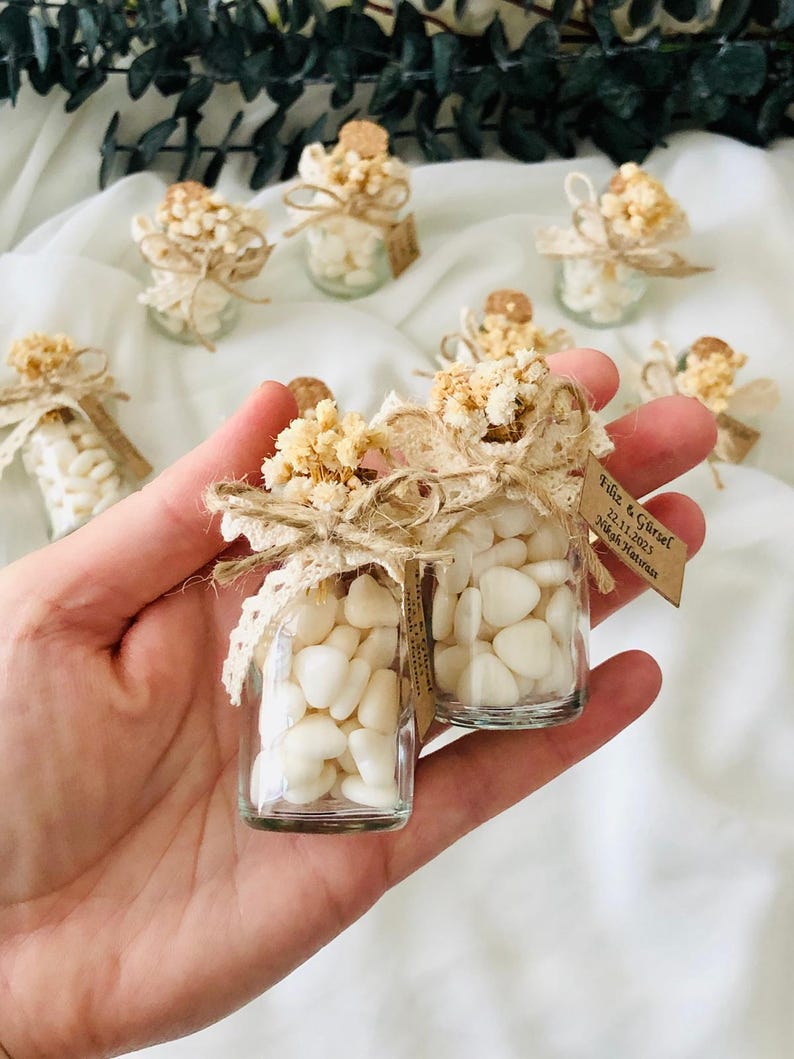 Peut inclure: Petits bocaux en verre remplis de bonbons blancs en forme de c&oelig;ur, orn&eacute;s de fleurs s&eacute;ch&eacute;es, de dentelle et de ficelle. Chaque bocal a un bouchon en li&egrave;ge et une petite &eacute;tiquette. Les bocaux sont dispos&eacute;s sur une surface blanche.