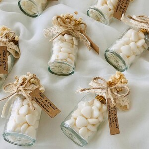 Peut inclure: Plusieurs petits flacons en verre remplis de bonbons blancs en forme de c&oelig;ur. Chaque bouteille est orn&eacute;e d'un n&oelig;ud d&eacute;coratif, de fleurs s&eacute;ch&eacute;es, de dentelle et d'une petite &eacute;tiquette avec du texte. Les bouteilles sont dispos&eacute;es sur une surface blanche.