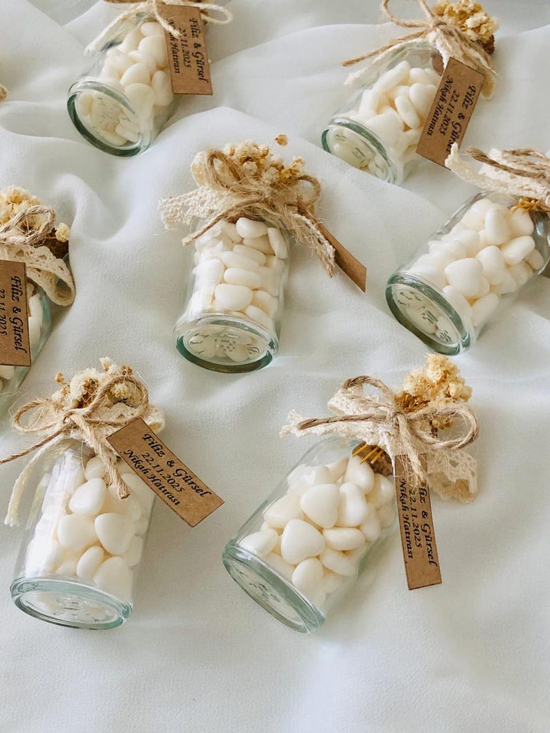 Peut inclure: Plusieurs petits flacons en verre remplis de bonbons blancs en forme de c&oelig;ur. Chaque bouteille est orn&eacute;e d'un n&oelig;ud d&eacute;coratif, de fleurs s&eacute;ch&eacute;es, de dentelle et d'une petite &eacute;tiquette avec du texte. Les bouteilles sont dispos&eacute;es sur une surface blanche.