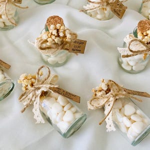 Peut inclure: Petites bouteilles en verre remplies de bonbons blancs en forme de c&oelig;ur, orn&eacute;es de fleurs s&eacute;ch&eacute;es, de dentelle et de ficelle. Chaque bouteille a un bouchon en li&egrave;ge et une petite &eacute;tiquette. Les bouteilles sont dispos&eacute;es sur un tissu blanc.