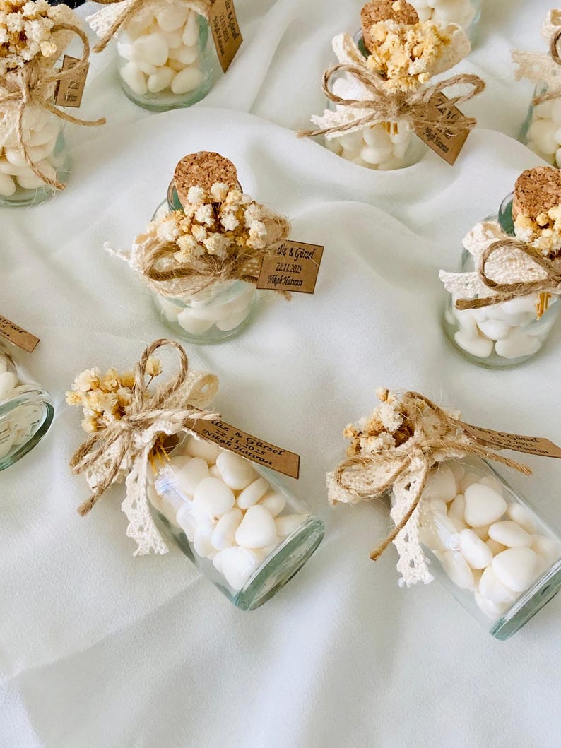 Peut inclure: Petites bouteilles en verre remplies de bonbons blancs en forme de c&oelig;ur, orn&eacute;es de fleurs s&eacute;ch&eacute;es, de dentelle et de ficelle. Chaque bouteille a un bouchon en li&egrave;ge et une petite &eacute;tiquette. Les bouteilles sont dispos&eacute;es sur un tissu blanc.