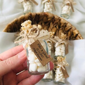 Peut inclure: Petites bouteilles en verre remplies de bonbons blancs, attach&eacute;es avec de la ficelle, de la dentelle et des fleurs s&eacute;ch&eacute;es. Chaque bouteille porte une petite &eacute;tiquette avec du texte. Les bouteilles sont dispos&eacute;es dans un panier tress&eacute;, pour un mariage ou un &eacute;v&eacute;nement.