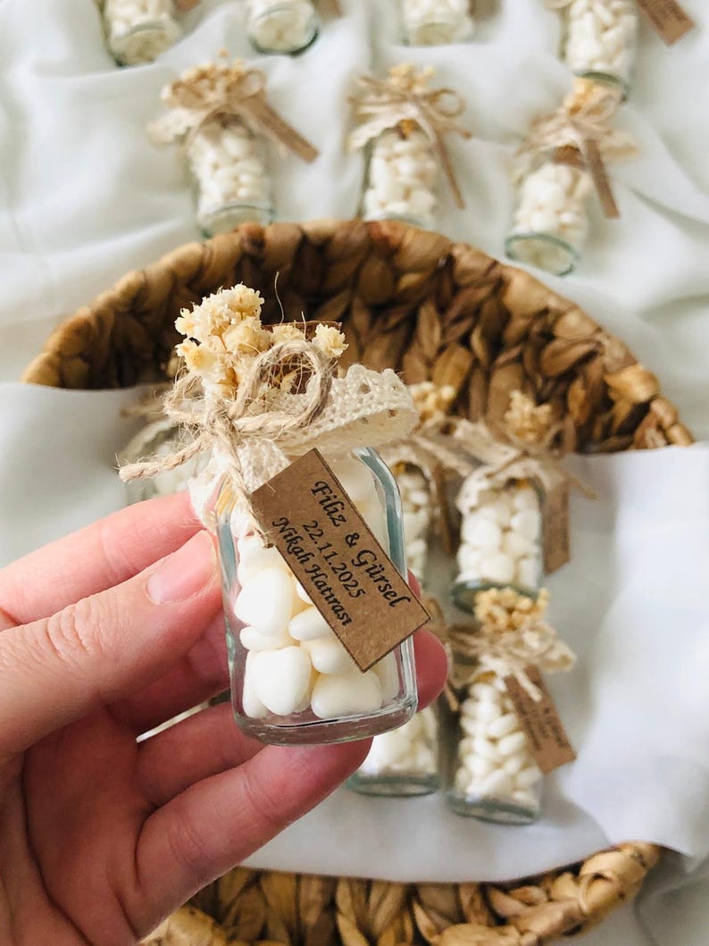 Peut inclure: Petites bouteilles en verre remplies de bonbons blancs, attach&eacute;es avec de la ficelle, de la dentelle et des fleurs s&eacute;ch&eacute;es. Chaque bouteille porte une petite &eacute;tiquette avec du texte. Les bouteilles sont dispos&eacute;es dans un panier tress&eacute;, pour un mariage ou un &eacute;v&eacute;nement.