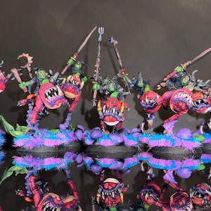May include: Miniature fantasy figures with green skin, red and pink bodies, and large teeth. The figures are mounted on bases with purple, blue, and pink fuzzy textures. They are holding weapons and are painted with detailed features.