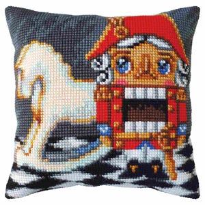 Puede incluir: Una almohada bordada colorida con un Cascanueces soldado con un caballo blanco en el fondo. El Cascanueces lleva un uniforme rojo y dorado con una banda azul. El caballo es blanco con una silla de montar y una brida azules.