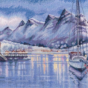 Puede incluir: Un paisaje montañoso nevado con un pequeño pueblo y un velero atracado en el puerto. El agua está tranquila y refleja las luces del pueblo.