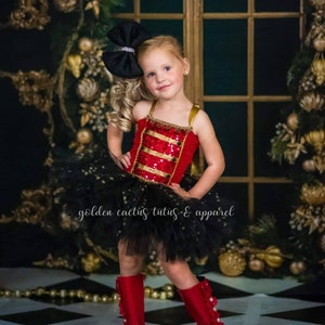 May include: A young girl wearing a red and gold sequined bodice with a black tulle tutu and red boots. The tutu has gold stars on it. The girl is wearing a black bow in her hair. The text "golden cactus tutus & apparel" is visible in the image.