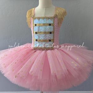 May include: Pink and gold tutu dress with a bodice featuring gold trim, sequins, and black buttons. The tutu skirt is layered pink tulle with gold star accents. The dress has gold fringe on the shoulders. The text "golden cactus tutus and apparel" is on the dress.