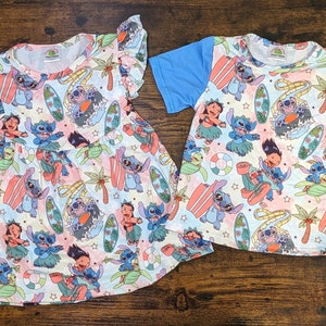 May include: A pink and blue dress and a blue and white shirt with a repeating pattern of Stitch from Lilo & Stitch surfing on a surfboard. The pattern also includes palm trees, stars, and a hula girl.