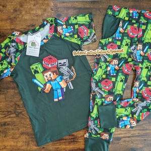 May include: Green Minecraft themed pajamas for kids. The shirt has a raglan sleeve design and features a graphic of Minecraft characters. The pants have a green background with a repeating pattern of Minecraft characters and blocks.