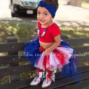 Könnte beinhalten: Ein junges Mädchen trägt ein rotes Crop-Top, einen blau-weißen Tutu und ein blaues Stirnband. Sie trägt weiße Cowboy-Stiefel mit rot-weiß-blauen Fransen. Der Tutu hat ein rot-weiß-blaues Farbschema. Das Mädchen sitzt auf einer Holzbank. Der Text "golden cactus tutus & apparel" ist im Bild sichtbar.