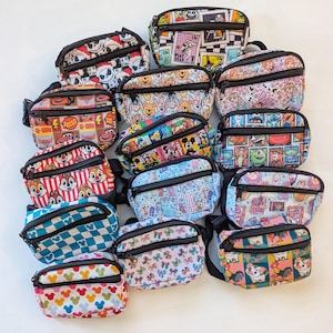May include: A collection of small, rectangular fanny packs with black zippers and adjustable straps. The packs feature various colorful patterns, including cartoon characters, checkered designs, and floral prints. The fanny packs are made of a durable material.