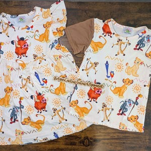 Pode incluir: Um vestido branco e uma camisa de manga curta com uma manga marrom, ambos com uma estampa de personagens do filme O Rei Leão. A estampa inclui Simba, Nala, Pumbaa, Timon e Zazu.
