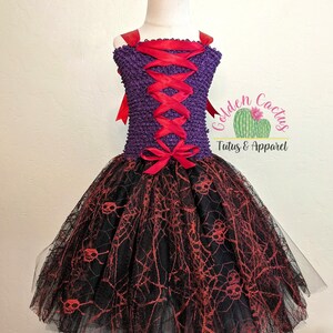 Könnte beinhalten: Lila und schwarzes Tüll-Tutu-Kleid mit roten Schleifenbändern und einer roten Schleife. Der Tüllrock hat ein Spinnennetz- und Totenkopf-Muster. Das Oberteil ist lila mit roter Schleifen-Schnürung. Das Kleid ist auf weißem Hintergrund. "Golden Cactus Tutus & Apparel" ist auf dem Kleid aufgedruckt.