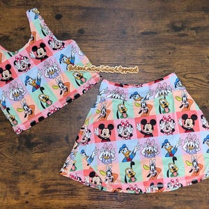 May include: A two-piece children's outfit featuring a tank top and skirt. The set is patterned with cartoon characters, including Mickey and Minnie Mouse, Donald Duck, and Pluto. The outfit has a colorful, pastel color scheme.