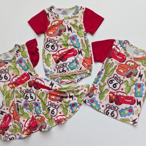 Puede incluir: Conjunto de ropa infantil con estampado de la Ruta 66 y Cars. El conjunto incluye un vestido, una camiseta de manga corta y un body. El patrón principal es un fondo claro con coches rojos, cactus y señales de la Ruta 66. Las mangas son de color rojo liso.