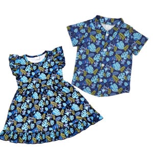 May include: A matching set of children's clothing featuring a blue dress and a button-down shirt. Both items are adorned with a tropical leaf and cartoon character pattern in shades of blue, green, and yellow. The dress has ruffled sleeves and a skirt.